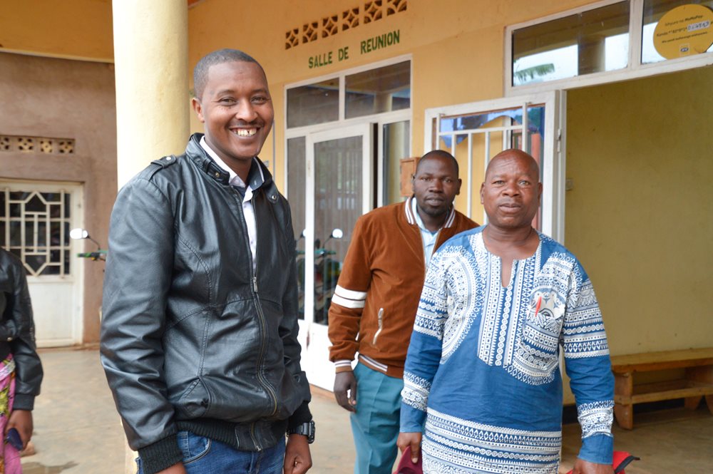 Three men smiling in front of yellow building with sign "Salle de Reunion"