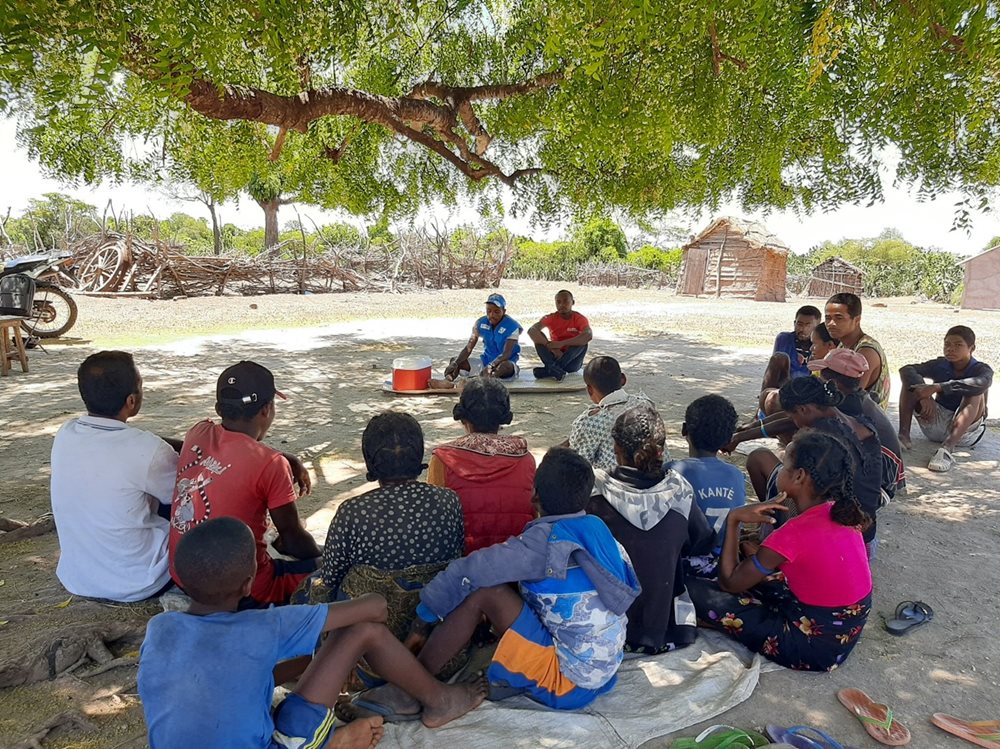 A group of people sit on the ground listening to two men speak.